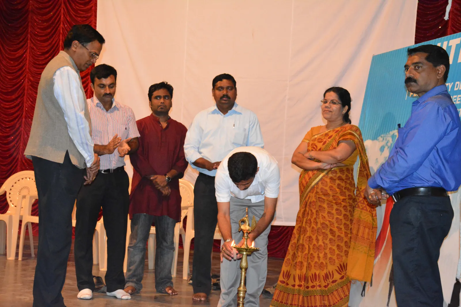People gathered on a stage lighting a traditional lamp in front of an event backdrop.
