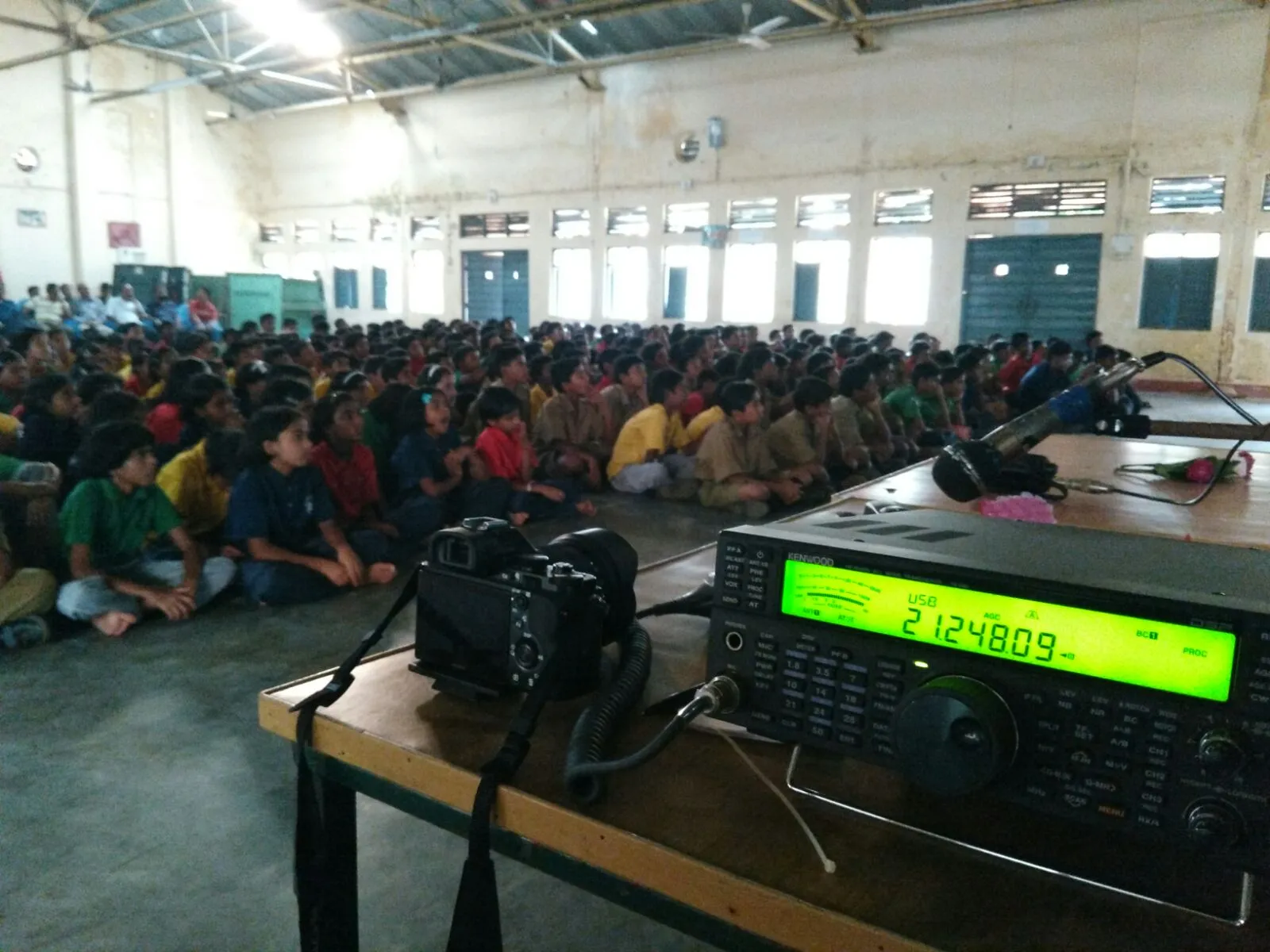 A radio transceiver and a camera on a table in a hall, with students seated on the floor.