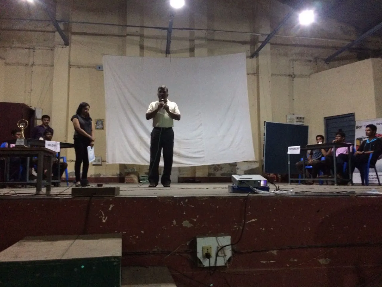 A speaker at a microphone on a stage, with students seated at tables on either side.